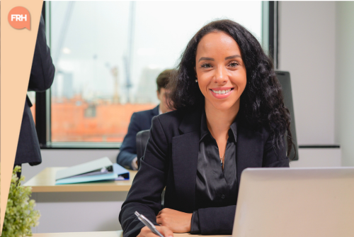 Jeune femme souriante en tenue professionnelle assise à un bureau avec un ordinateur, dans un environnement de travail moderne.