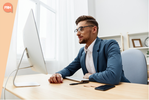 Jeune homme en costume assis à son bureau, concentré sur son écran d’ordinateur dans un bureau lumineux et épuré.
