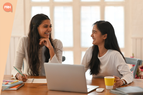 Deux jeunes femmes souriantes travaillant ensemble à une table, échangeant des idées devant un ordinateur portable dans un espace lumineux.