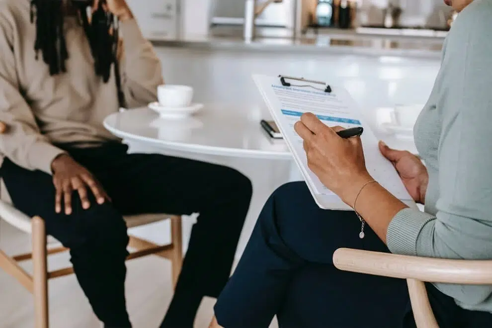 Une personne assise prend des notes sur un clipboard face à quelqu’un assis à une table ronde.